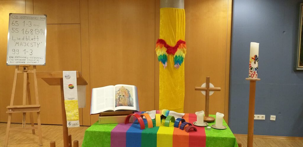Das Bild zeigt einen bunt in Regenbogenfarben geschmückten Altar mit aufgeschlagener Bibel, hellem Holzkreuz und Osterkerze.